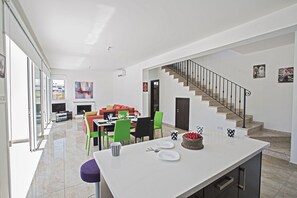 A modern kitchen with a central island, dining area with green chairs, and a staircase leading to an upper level.