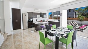 Una cocina moderna con armarios oscuros y electrodomésticos de acero inoxidable, un comedor con sillas verdes y un patio con sillones y vistas al exterior.
