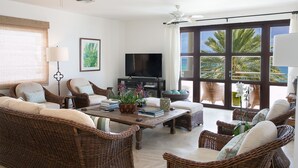 A spacious living room with wicker furniture, a flat-screen TV, and large windows offering a view of palm trees and the ocean. - Blue Dove | Beach Front - Located in  Beautiful Cupecoy with Private Pool (Cupecoy)