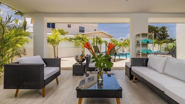 Ein moderner Außenbereich zum Sitzen mit Korbmöbeln, einem Glastisch und Blick auf einen Pool sowie Gebäude im Hintergrund.
