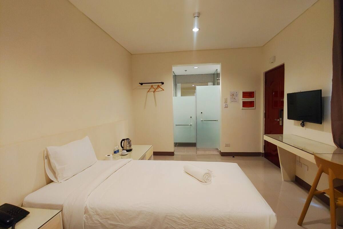 A hotel room with a bed, a desk with a TV, a telephone, and a kettle.