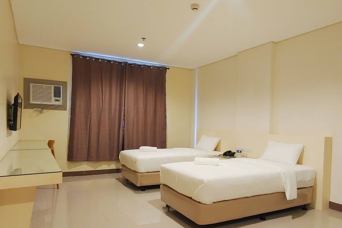 A hotel room with two beds, a desk, a chair, a window with curtains, and a wall-mounted air conditioner.