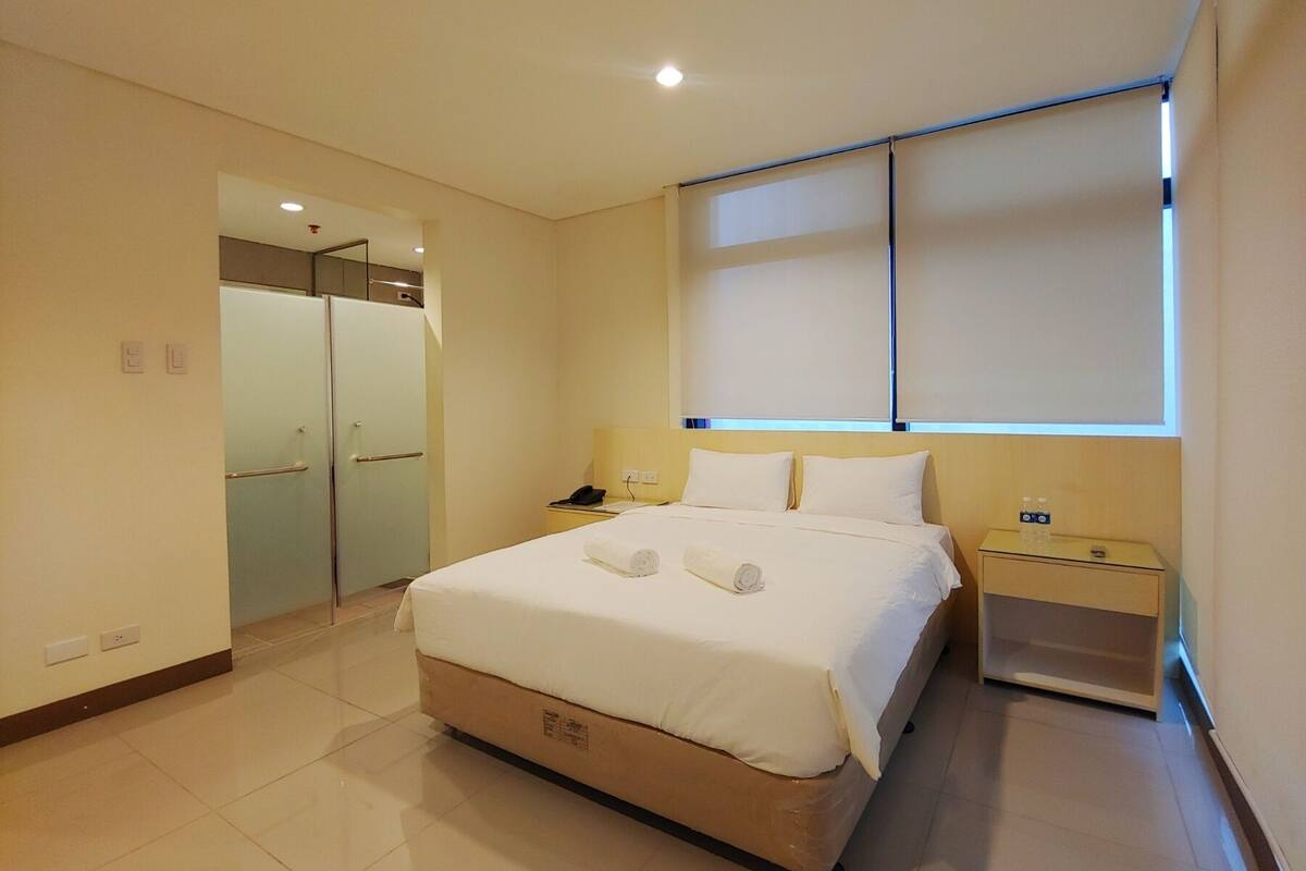 A hotel room with a bed, bedside table, and a window with blinds.