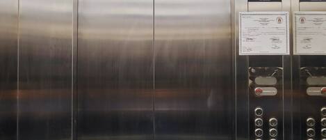 An elevator interior with stainless steel doors, a digital display, emergency call button, and a no-smoking sign.