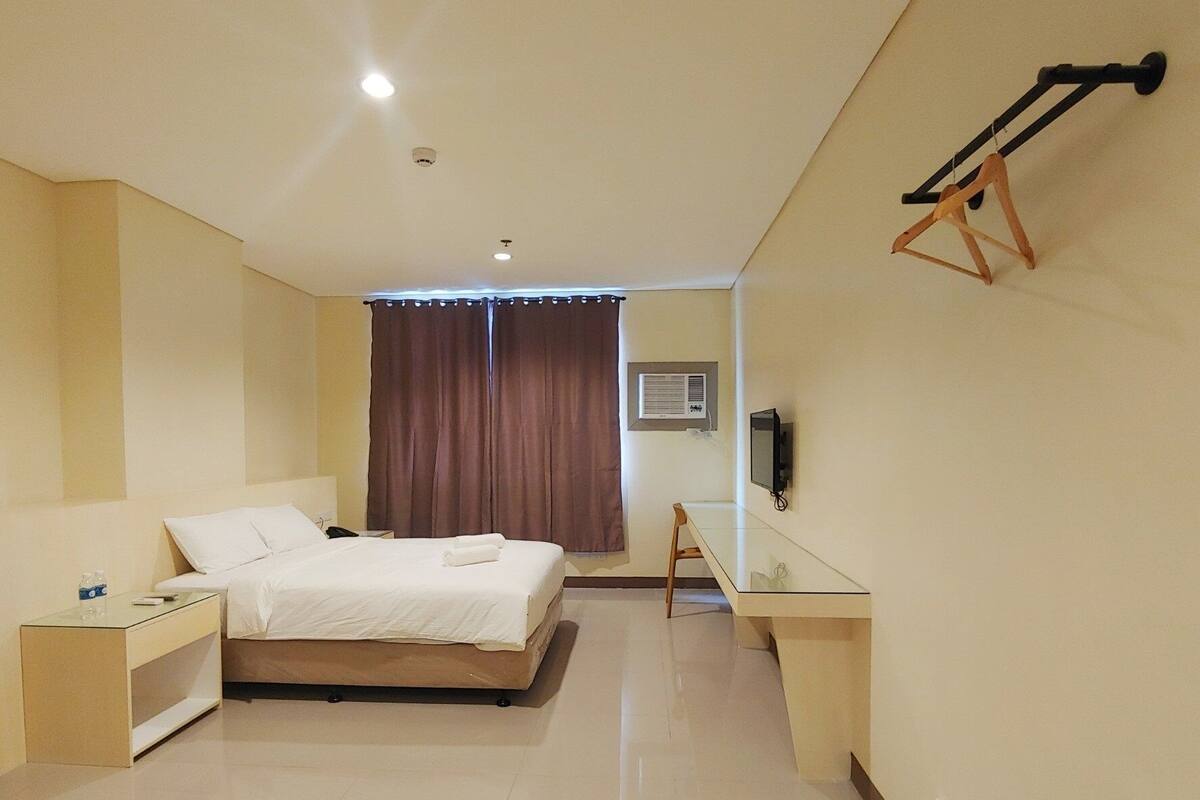 A hotel room with a bed, bedside table, a desk with a chair, an air conditioning unit, and a TV mounted on the wall.