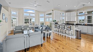 A spacious living area with a grey sofa set, a coffee table, and a dining area with bar stools. The kitchen features white cabinets and a black countertop.
