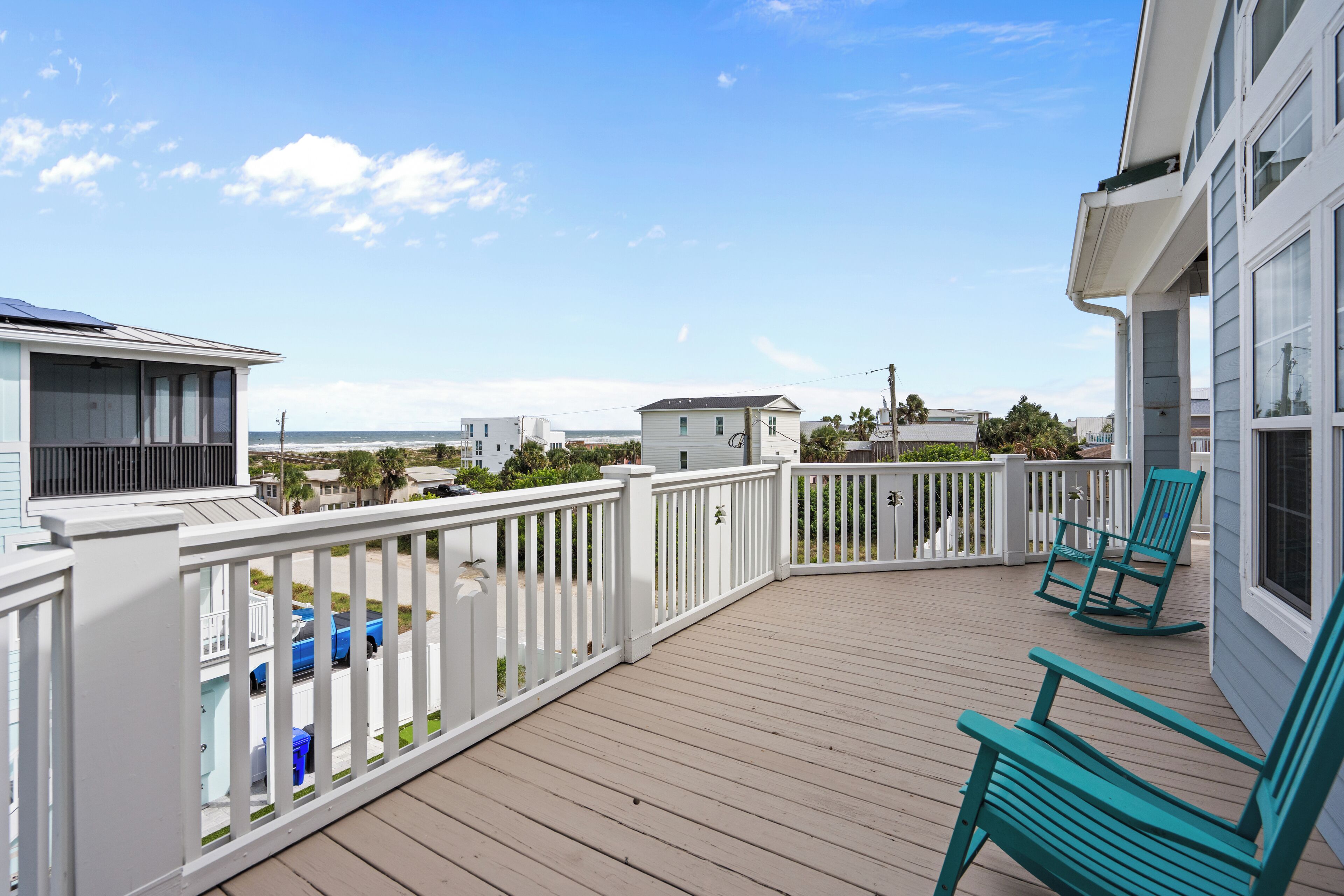 A wooden deck with white railings, two green rocking chairs, and a view of the ocean and other buildings.