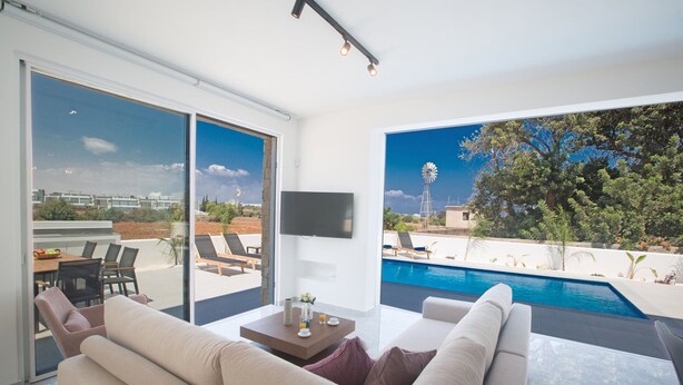 Un salon moderne doté d’une grande porte vitrée donnant sur une piscine, d’un téléviseur à écran plat et d’un coin repas avec une table et des chaises.