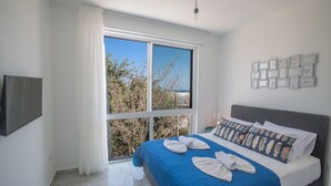 Une chambre d’hôtel dotée d’une grande fenêtre, d’un lit avec une literie bleue et des coussins décoratifs, d’un téléviseur à écran plat fixé au mur et d’une série de tableaux encadrés accrochés au mur.