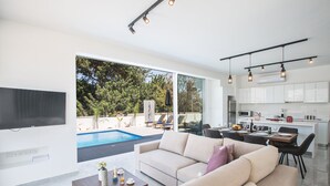 Un salon moderne avec un grand canapé, une table basse et un coin repas avec une table dressée pour deux personnes. On peut apercevoir une piscine à travers les portes vitrées, et la cuisine est équipée de meubles blancs et d’un four à micro-ondes.