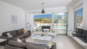 Un salon moderne avec un ensemble de canapés, une table basse et un téléviseur. Il y a un balcon avec une piscine et du mobilier d’extérieur.