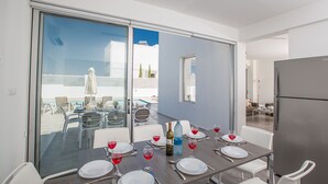 A dining area with a table set for four, a pool visible through the glass doors, and a modern kitchen with a refrigerator. - The Complete Guide to Renting Your Exclusive Holiday Villa in Protaras with Private Pool and Close to the Beach (Protaras)