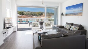A modern living room with a grey sofa, a coffee table, and a TV on a stand. There is a large window with a view of a pool and outdoor furniture. - Protaras Holiday Villa with Private Pool, Protaras Villa 1554 (Protaras)