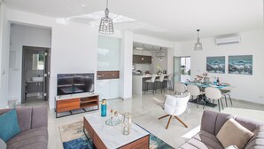 Un salon moderne avec un téléviseur, un canapé et une table basse. On y trouve également un coin repas avec des chaises et une table. La cuisine est visible au fond.