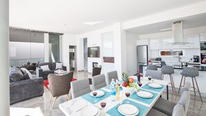 A modern living space with a dining area, grey sofas, and a kitchen with white cabinets.