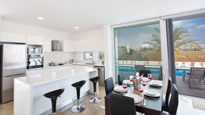 A modern kitchen with white cabinetry, a built-in oven, and a microwave. There is a dining area with a table set for two, two black bar stools, and a view of a swimming pool and palm trees outside.