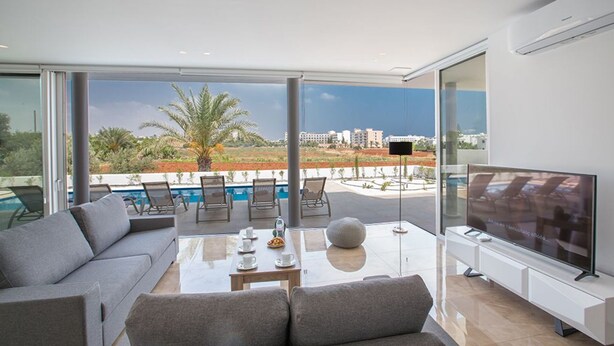 A modern living room with a sofa, a coffee table, and a TV. There is a balcony with a pool and palm trees.