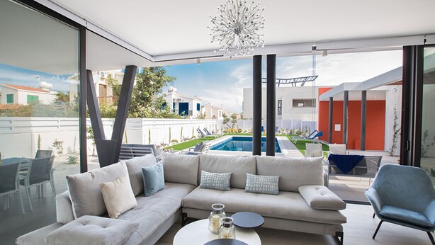 A modern living room with a large sofa, a coffee table, and a view of a swimming pool and outdoor area. - Your Dream Holiday Villa with Private Pool in Protaras most Exclusive Neighbourhood (Protaras)