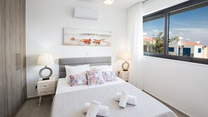A modern bedroom with a bed, two bedside tables, a round mirror, a ceiling light, a window with a view of buildings, and a wall-mounted air conditioner. - Your Dream Holiday Villa with Private Pool in Protaras most Exclusive Neighbourhood (Protaras)