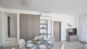 A modern dining area with a round table set for four, surrounded by white chairs. There are wooden cabinets and a television on a stand to the right. - Your Dream Holiday Villa with Private Pool in Protaras most Exclusive Neighbourhood (Protaras)