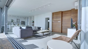Een moderne woonkamer met een bank, salontafel en een fauteuil. Achtergrond toont een keuken met witte kasten en een eethoek.