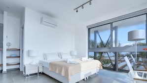 Een moderne hotelkamer met een groot bed, twee nachtkastjes, een bureau en een stoel. Er is een groot raam met uitzicht op palmbomen en een gebouw.