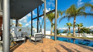 Een modern buitenzwembad met een houten terras, ligbedden en uitzicht op palmbomen en een berg.