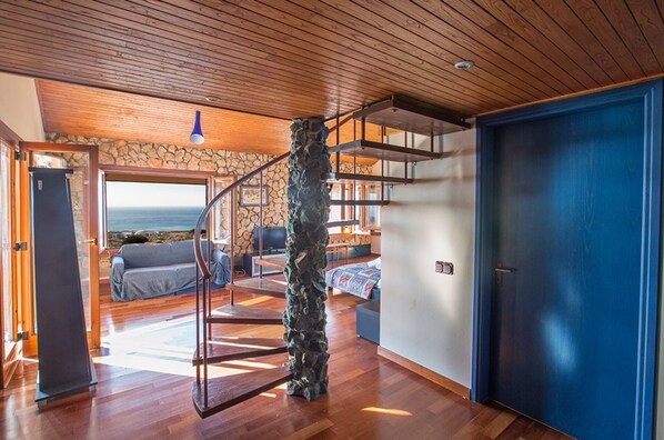Amplia sala de estar con piso de madera, un pilar de piedra, una puerta azul y vista al océano a través de un ventanal grande.
