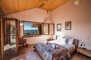 A bedroom with a large bed, a wooden ceiling, a view of the sea, a dresser, a nightstand, and a mirror.