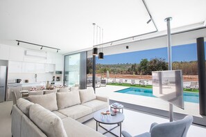 A modern living room with a sofa, a coffee table, and a TV. There is a kitchen area with white cabinets and a dining table. A pool is visible through large windows. - The Ultimate 5 Star Holiday Villa in Protaras with Private Pool and Close to the Beach (Protaras)