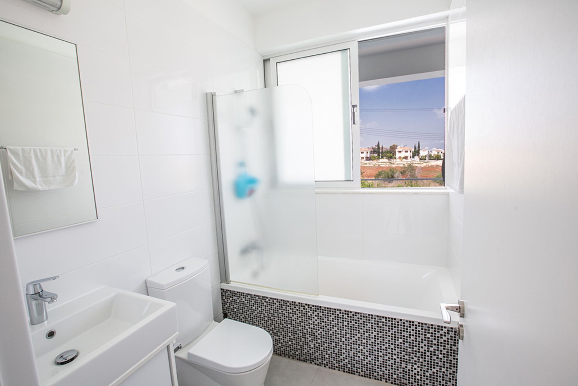 Une salle de bain moderne dotée d’une douche à parois de verre, de toilettes, d’un lavabo et d’une fenêtre donnant sur les bâtiments environnants.
