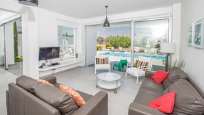 Un salon moderne avec un canapé, une table basse et un téléviseur. On peut apercevoir une piscine à travers les grandes fenêtres.