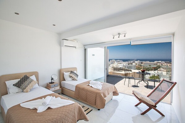Ein modernes Hotelzimmer mit zwei Betten, einem Balkon mit Blick auf Stadt und Meer sowie einer Sitzecke.