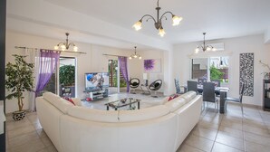 A spacious living room with a white sofa, a coffee table, and a TV. There are two chairs, a dining table, and a chandelier. - The Complete Guide to Renting Your Exclusive Holiday Villa in Protaras with Private Pool and Close to the Beach (Protaras)