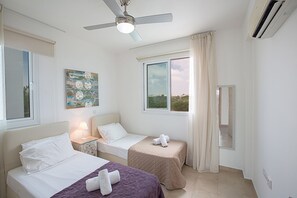 A hotel room with two beds, a ceiling fan, a painting, and a view of the landscape through the windows. - Rent Your Dream Protaras Holiday Villa and Look Forward to Relaxing Beside Your Private Pool (Paralimni)