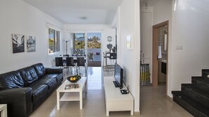 Ett modernt vardagsrum med en svart lädersoffa, ett vitt soffbord och en tv på ett vitt tv-bord. Det finns en matplats med ett bord och stolar, och ett kök som syns genom en öppen dörr.