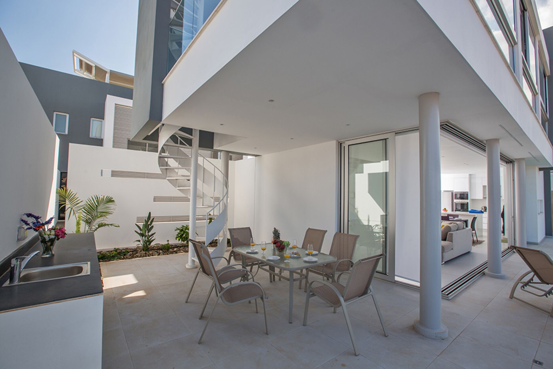A modern outdoor dining area with a glass table and chairs, a staircase, and a view of the interior kitchen.