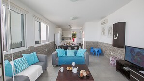 A compact living space with a sofa, a coffee table, and a television. There is a kitchen area in the background with a dining table and chairs.