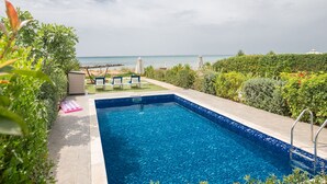 A rectangular swimming pool with a view of the ocean, surrounded by greenery and a few lounge chairs.
