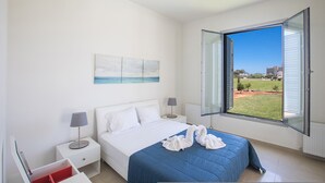 A bedroom with a bed, two bedside tables, a nightstand, a window with an open view, and a painting on the wall.