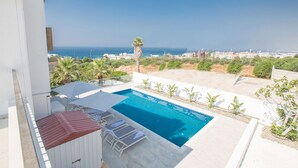 Una piscina en la azotea con agua cristalina, rodeada de sillones y sombrillas, con vistas a un paisaje costero y al mar, y edificios a lo lejos.