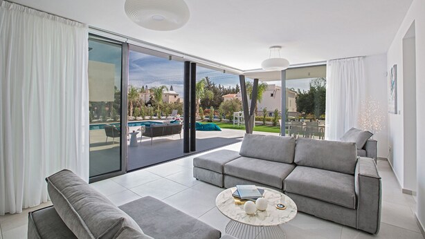 A spacious living area with a grey sofa set, a round coffee table, and a large sliding glass door leading to an outdoor pool area. - The Complete Guide to Renting Your Exclusive Holiday Villa in Protaras with Private Pool and Close to the Beach (Protaras)