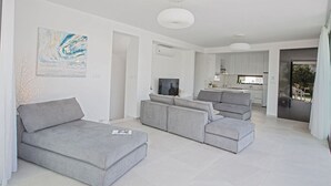 A modern living room with grey sofas, a flat-screen TV, and a kitchen in the background. - The Complete Guide to Renting Your Exclusive Holiday Villa in Protaras with Private Pool and Close to the Beach (Protaras)