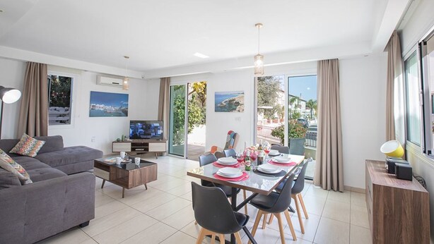 A modern living room with a sofa, a coffee table, a TV, and a dining area with chairs and a table set for two.
