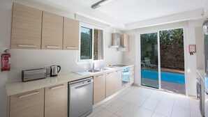 A modern kitchen with a dishwasher, a kettle, and a toaster. There is a window providing natural light and a view of a swimming pool outside.