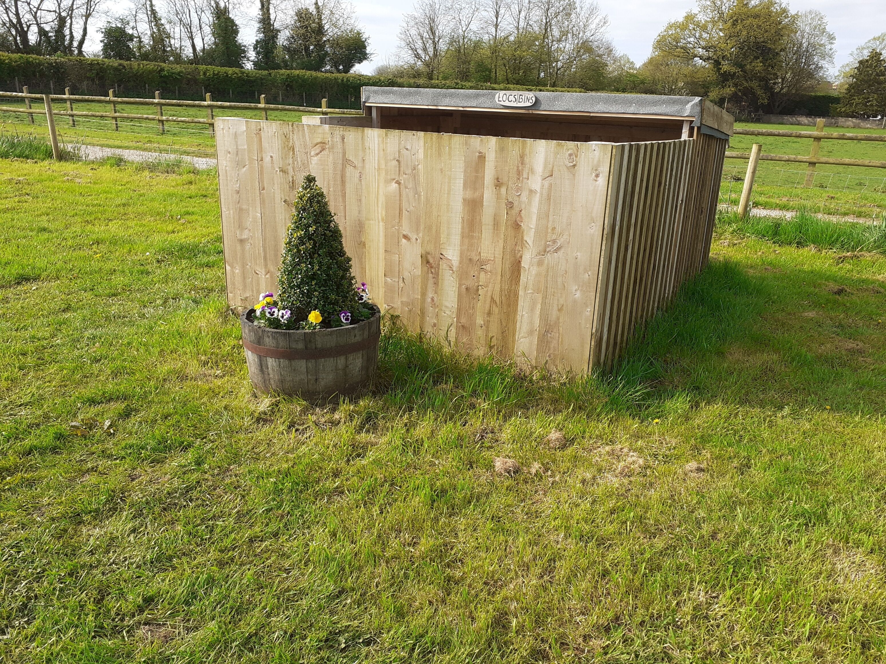 Een houten tuinhuisje met een bord waarop ‘LOGS BINS’ staat, en een kleine bloemenplanter met bloemen.