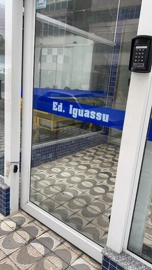 A glass door with a keypad lock, a tiled floor, and a blue banner with "Ed. Iguassu" text. - Loft Vista Mar (Guarapari)