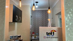 A reception area with a counter displaying the logo 'LoFi Vista' and a social media handle '@loft.guarapari'. There are light fixtures hanging from the ceiling and a patterned wall in the background. - Loft Vista Mar (Guarapari)