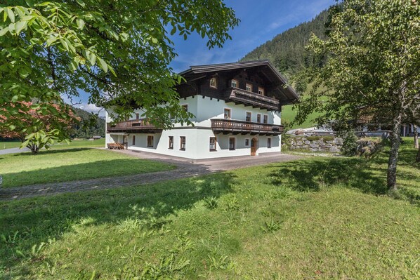 Un bâtiment de style chalet traditionnel, doté d’un balcon, entouré d’une végétation luxuriante et avec des montagnes en toile de fond.