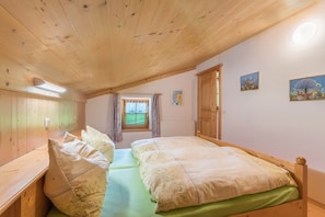 A room with wooden ceiling and walls, a bed with green bedding, a bench, a window with curtains, and a radiator.
